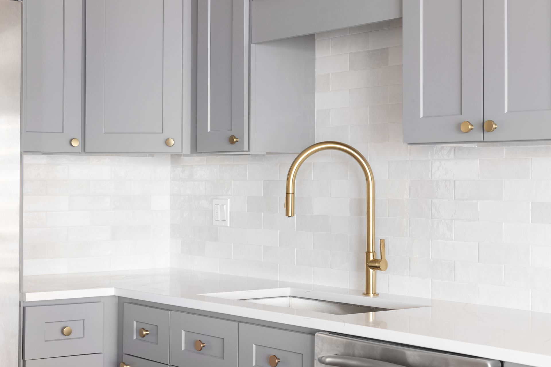 Gray kitchen cabinets, white backsplash, gold faucet, white countertop.