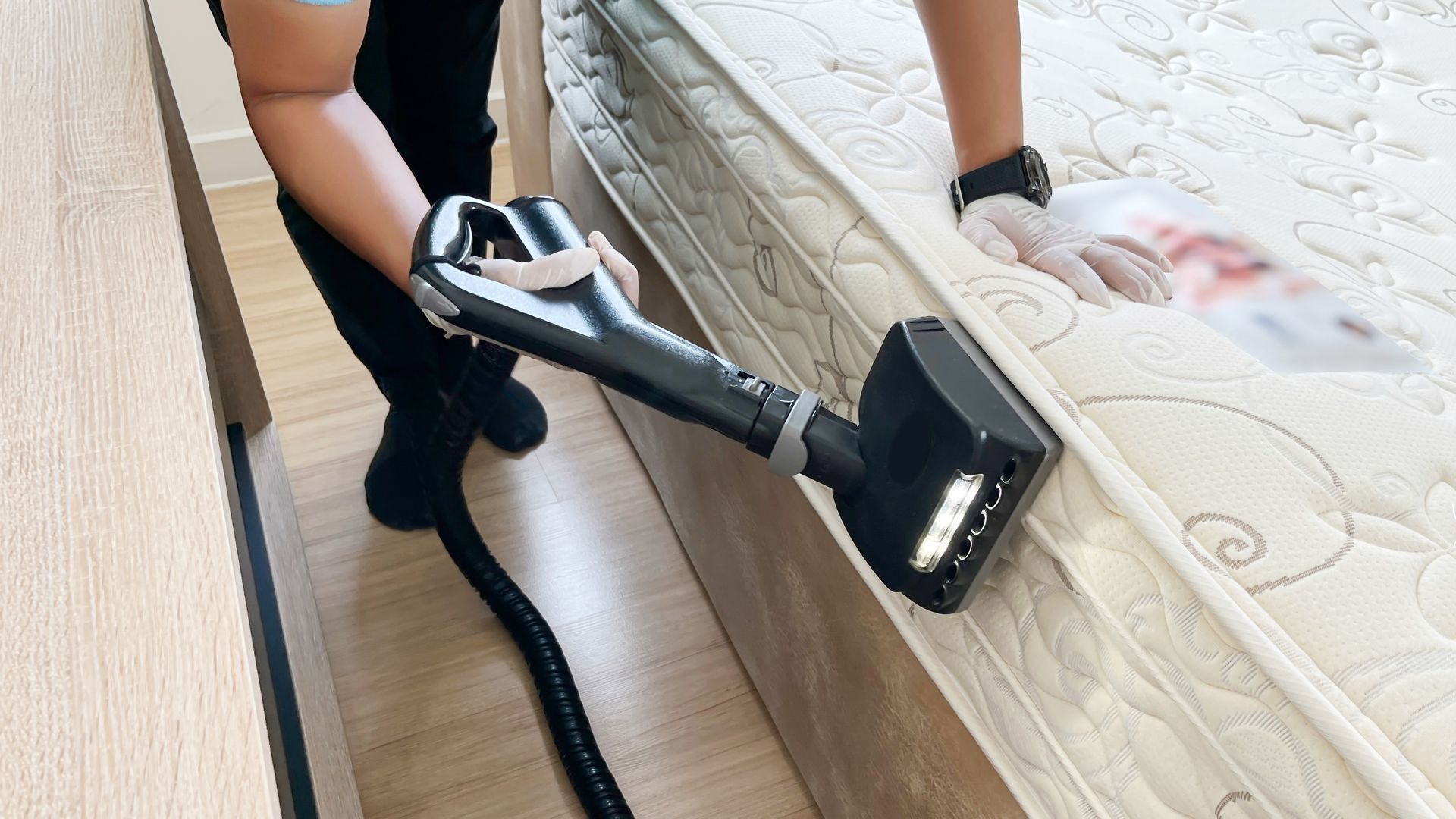 Person vacuuming a mattress, wearing black pants and socks, with a black vacuum and white gloves.