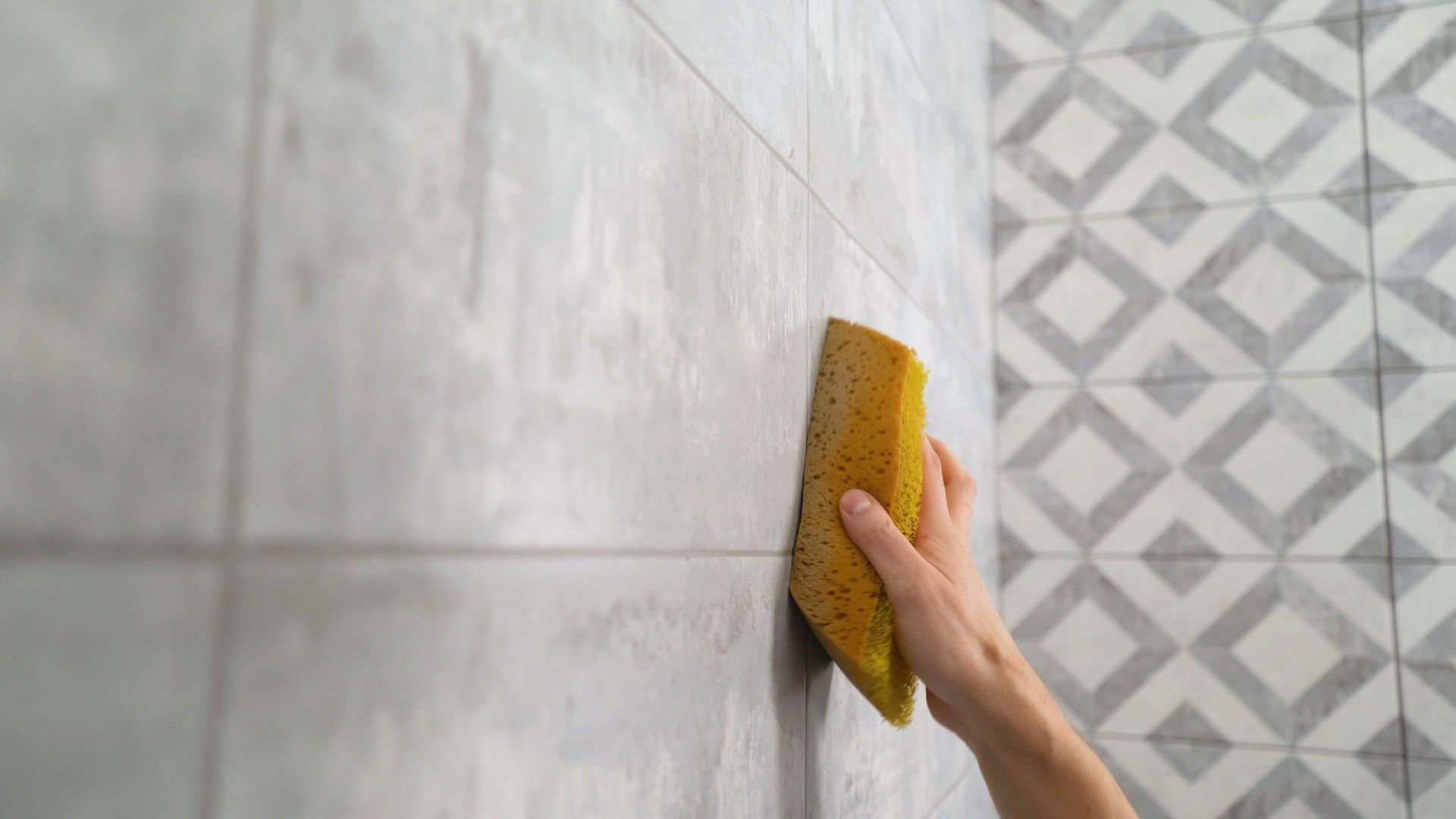 Hand wiping a light gray tiled wall with a yellow sponge, beside a patterned tile section.