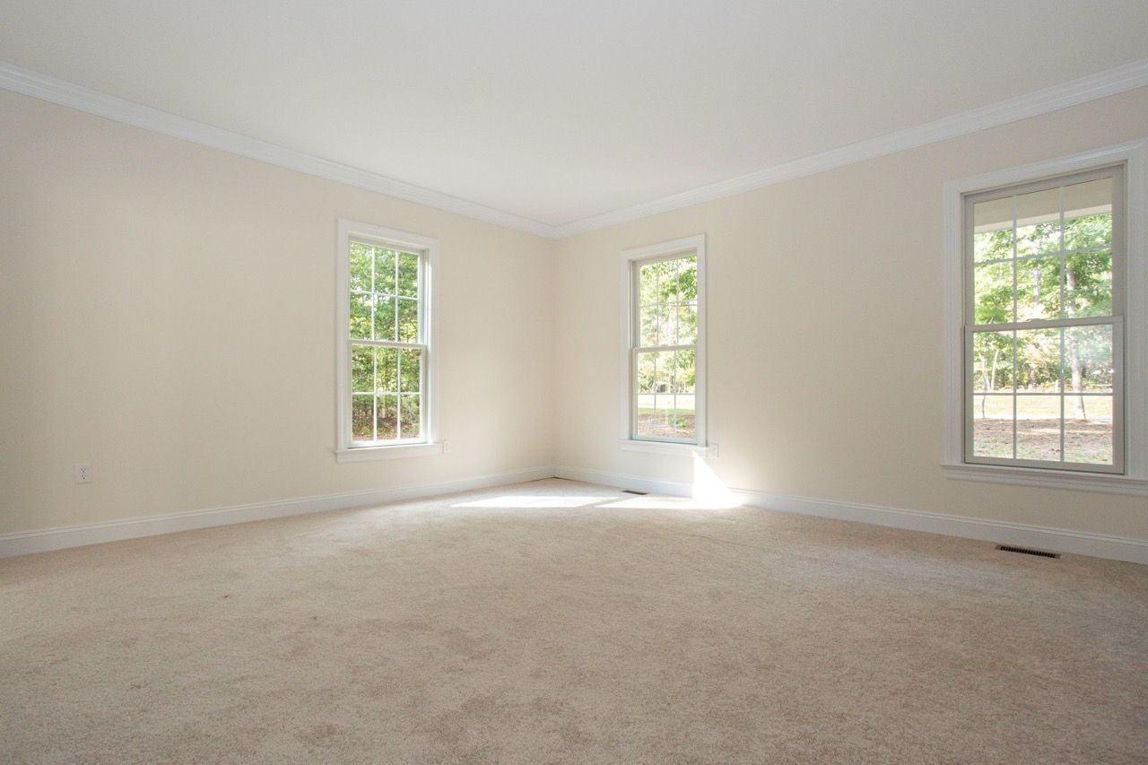 Empty beige carpeted room with three tall windows and white trim, filled with natural light