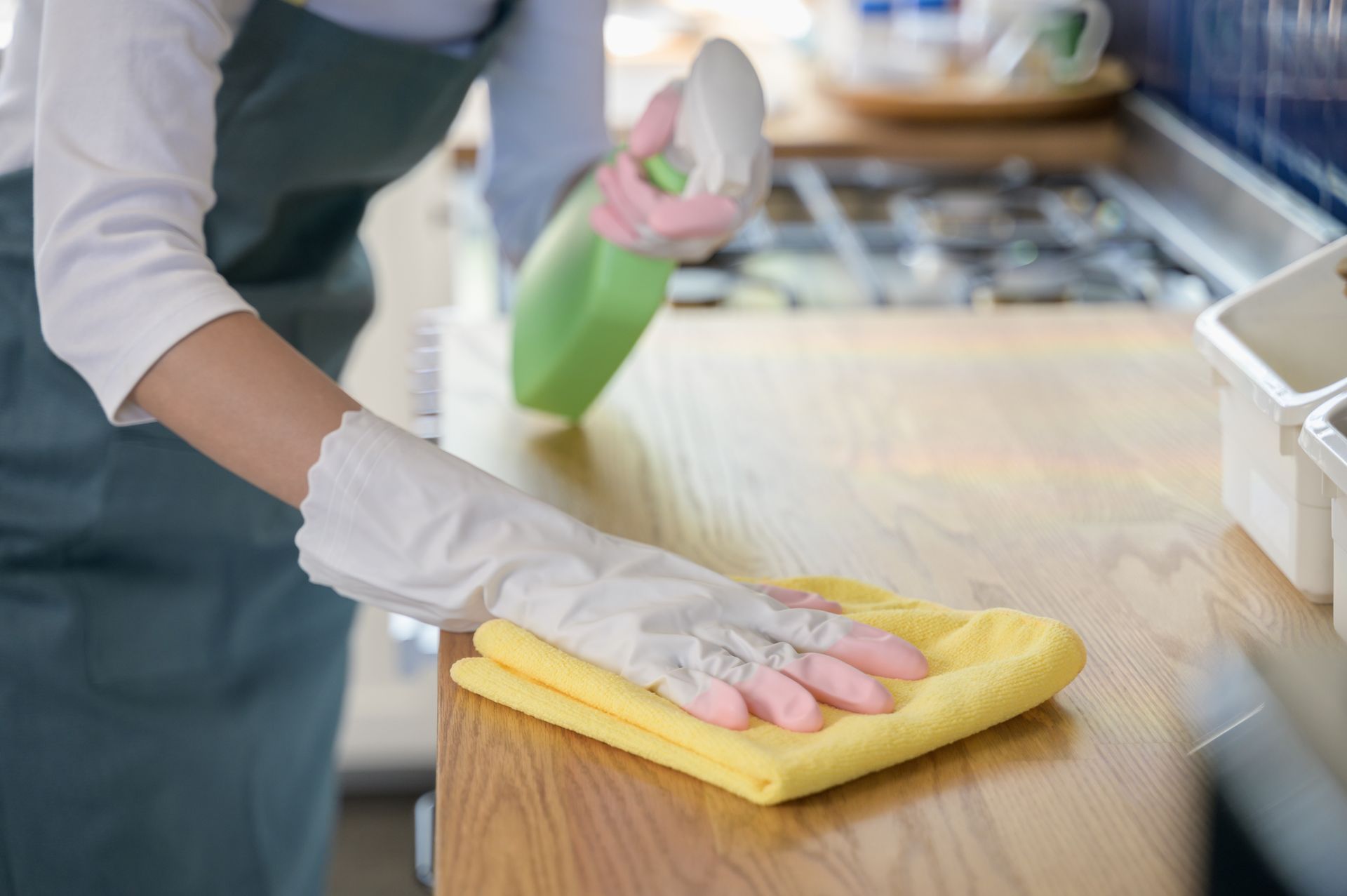 Person wearing gloves wiping a wooden countertop with a yellow cloth and spray bottle in a kitchen.