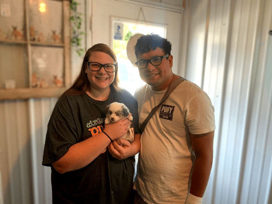 Two people smiling indoors, holding a small puppy near a window