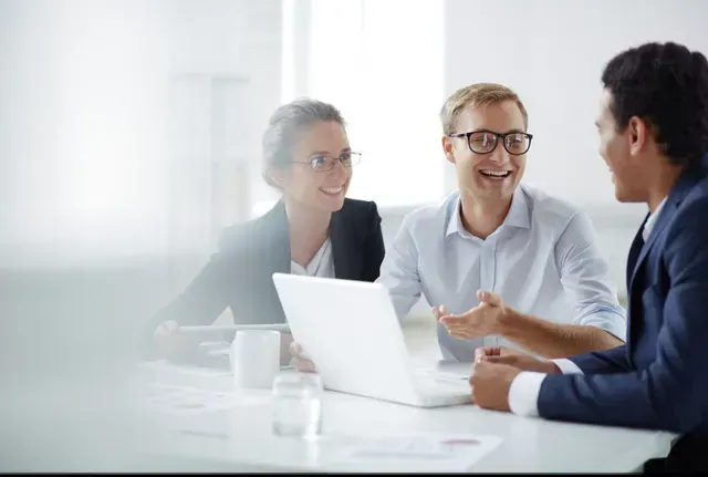 Three people in a bright office meeting around a laptop, smiling and talking.