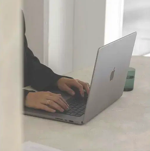 Hands typing on a silver laptop at a bright desk with a green mug nearby.
