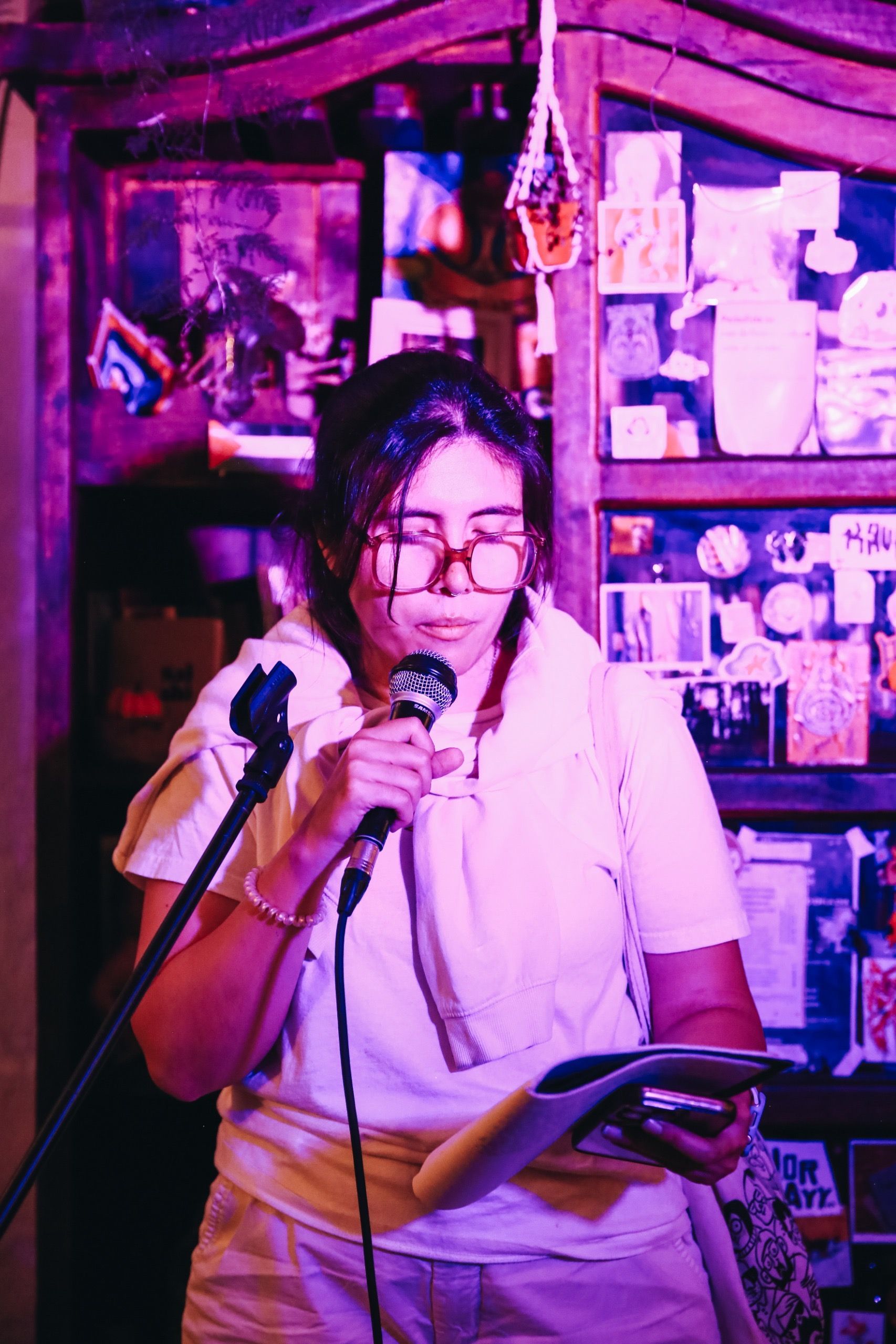 Una mujer con gafas lee un papel frente a un micrófono, parada frente a un estante con diversos artículos y un tono púrpura.