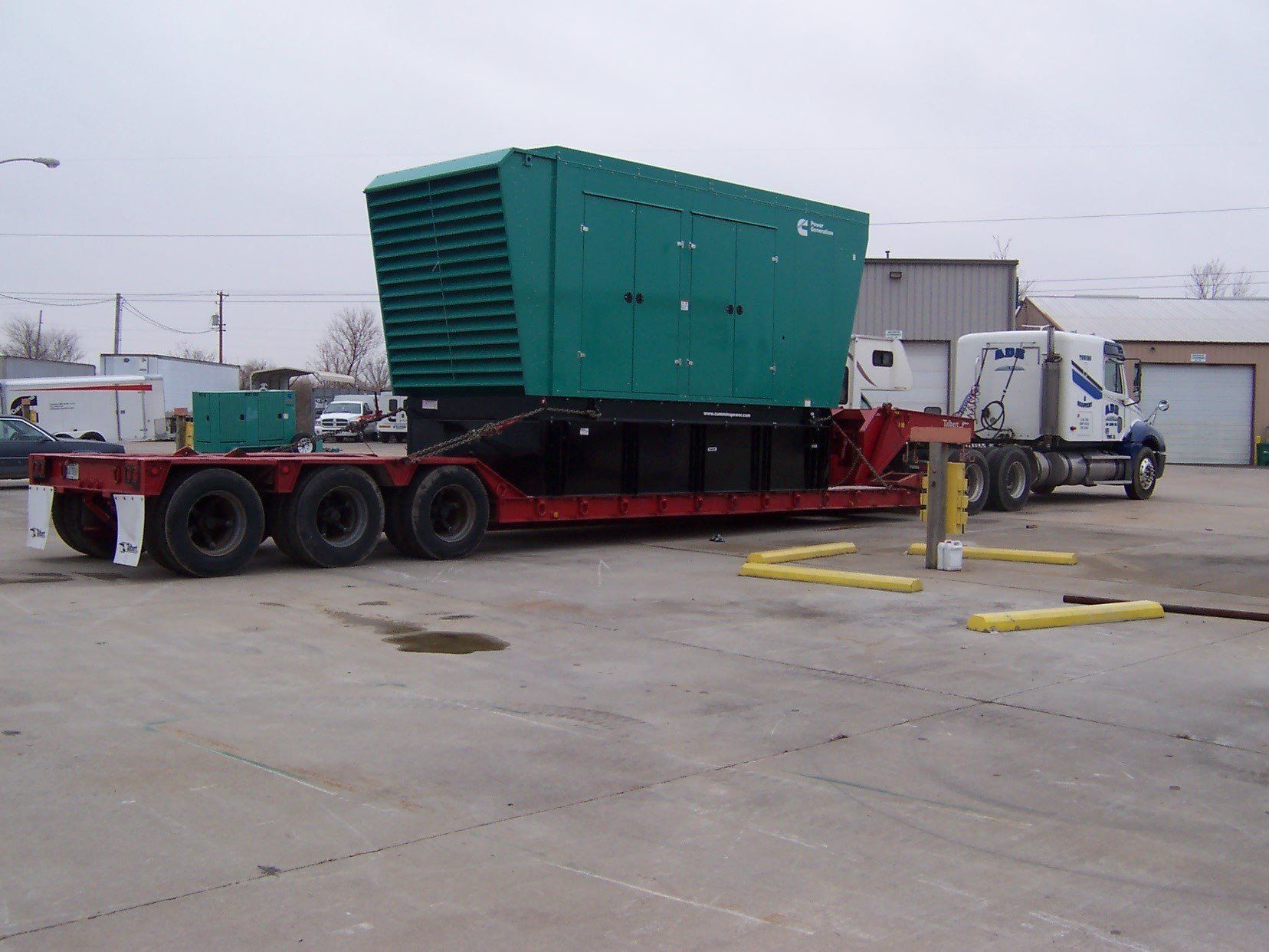 Green industrial machine on a red trailer in a concrete yard, with a white truck nearby.