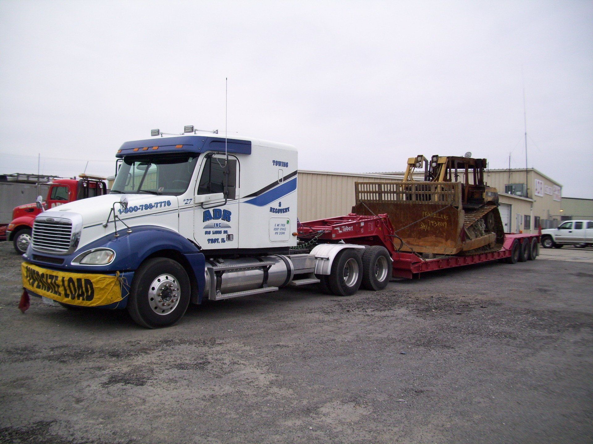 White and blue semi-truck hauling a red flatbed trailer with a large rusted machine on a gravel lot