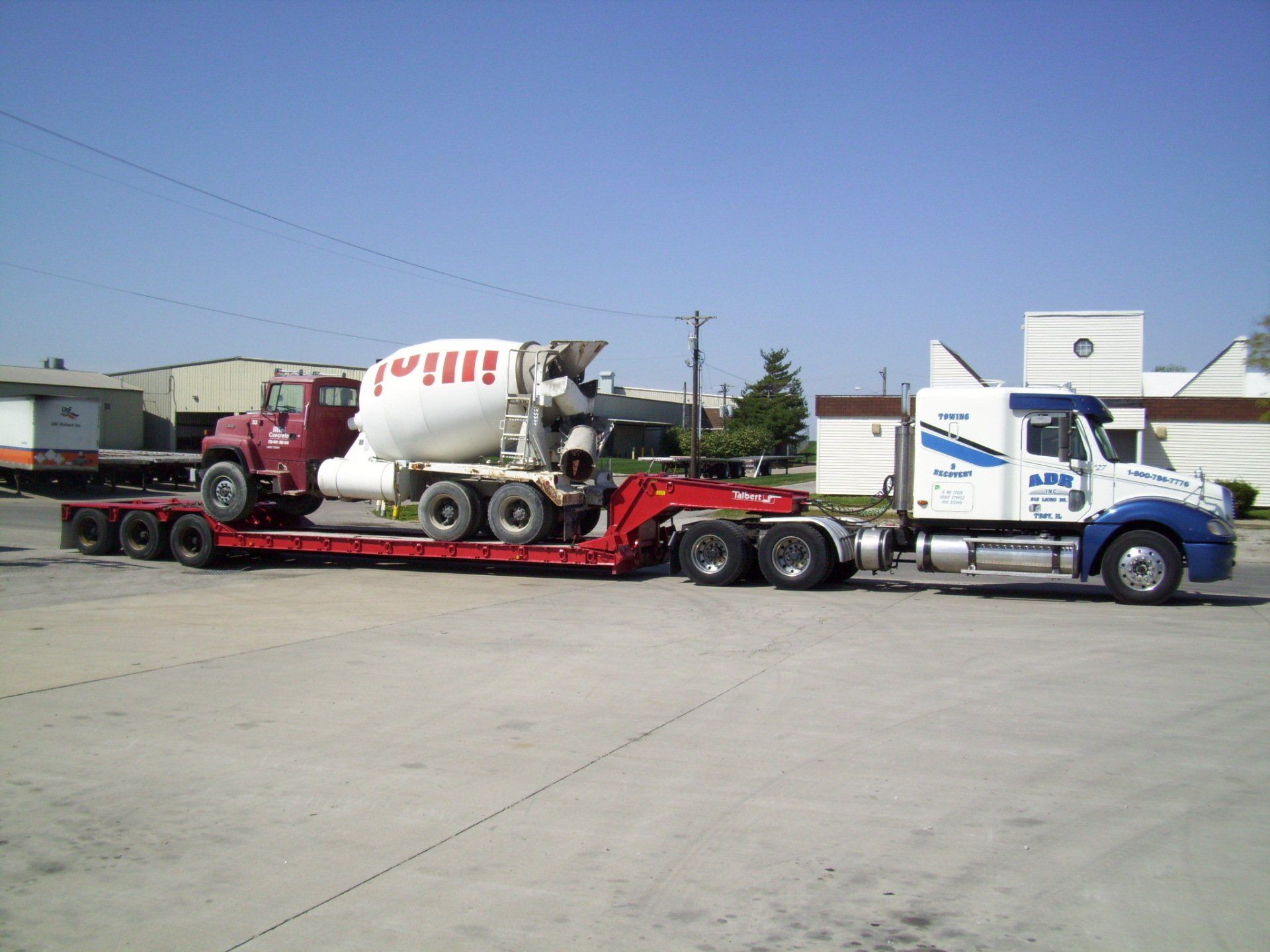 Blue-and-white semi truck hauling a red flatbed with a cement mixer in an open lot