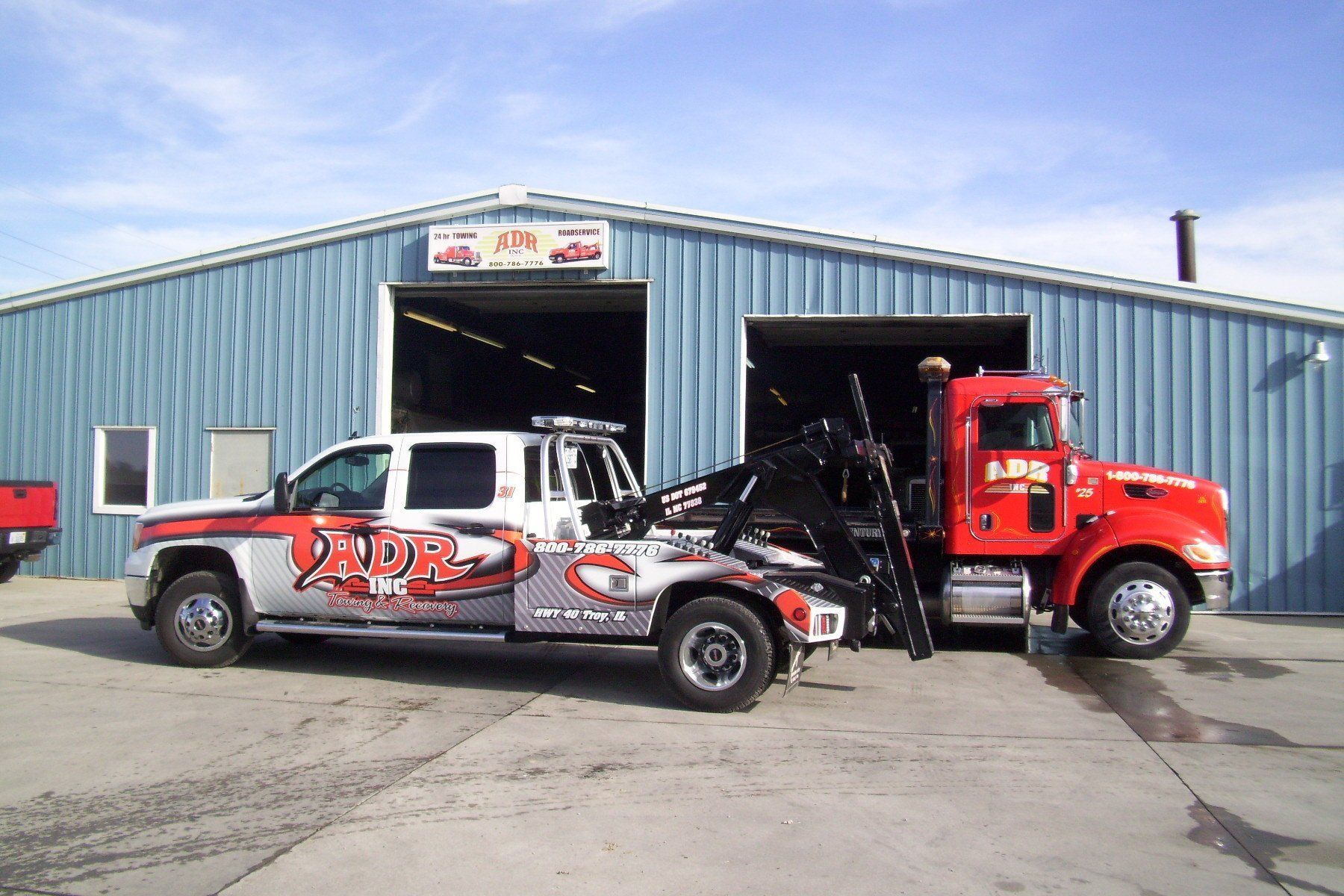 Tow truck and silver pickup with graphics parked outside a blue auto repair garage