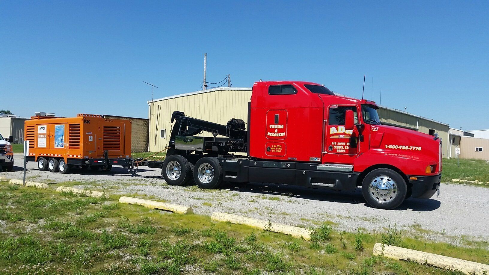 Red semi-truck hauling an orange trailer parked on a gravel lot under a clear blue sky