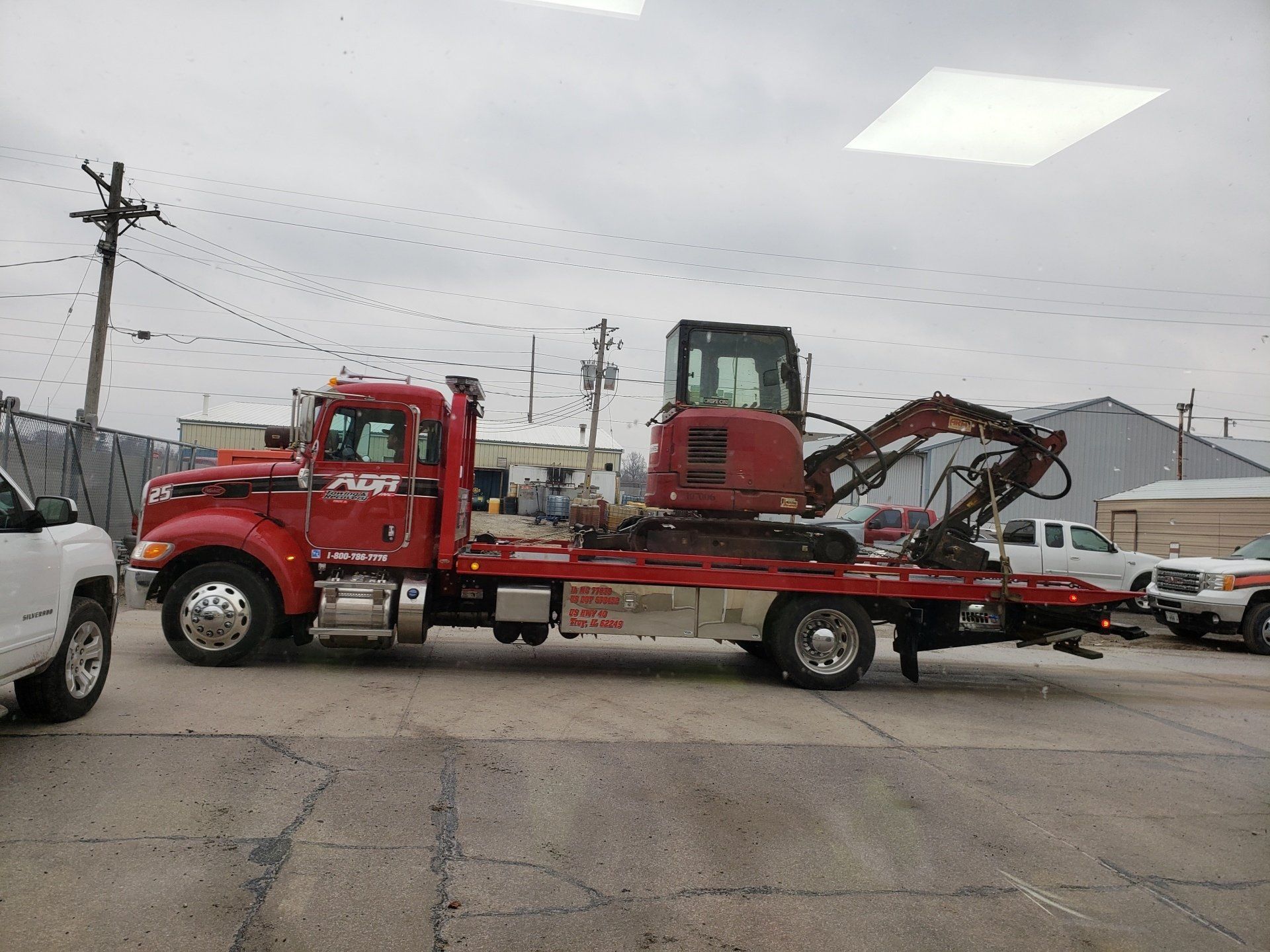 Red flatbed tow truck hauling a red excavator on a city street under cloudy skies