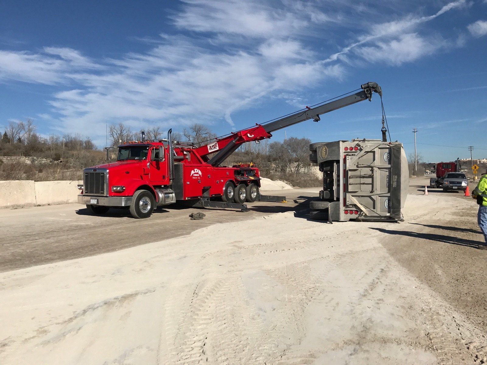 Red tow truck lifting an overturned gray vehicle on a dirt roadside under a blue sky