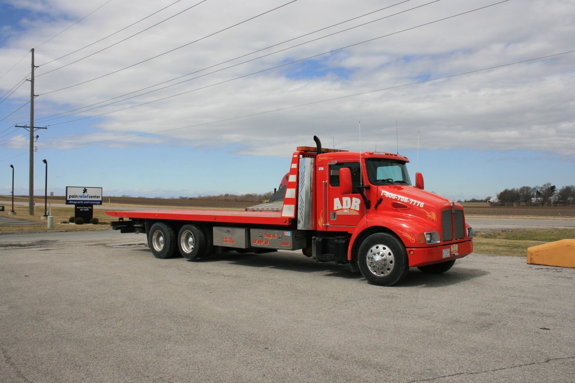 Red flatbed tow truck parked on a gravel lot under a cloudy sky