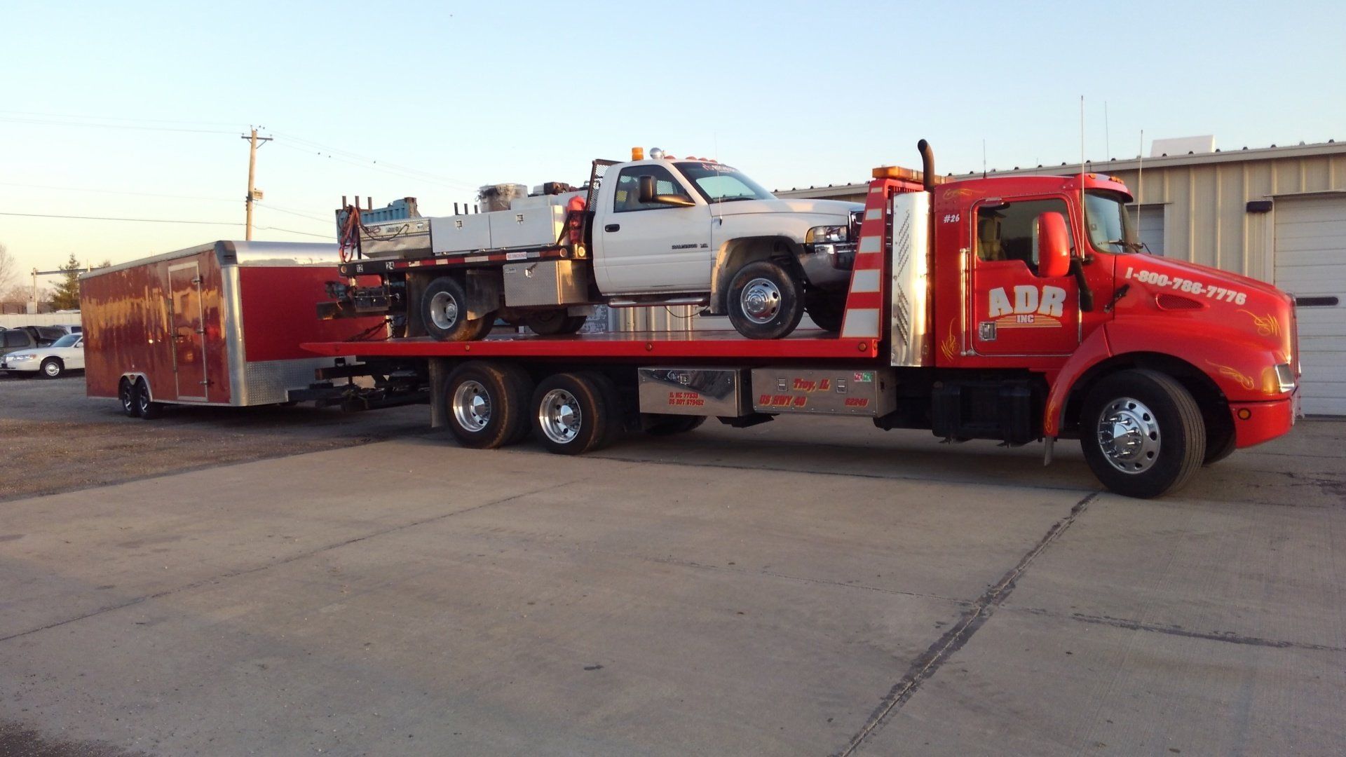 Red tow truck hauling a silver pickup and trailer on a road at sunset