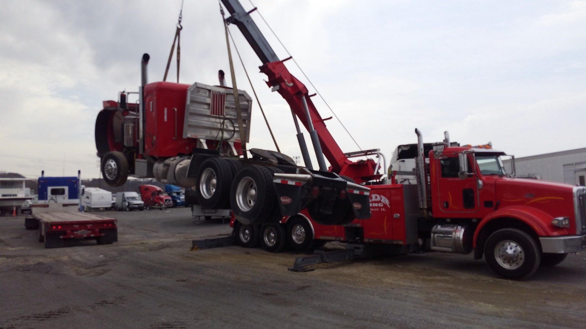 Red tow truck lifting a red semi-truck cab in an industrial lot