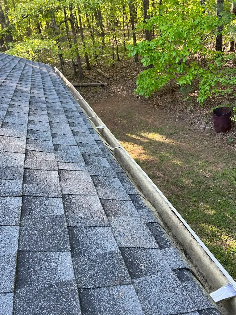 Rooftop with gray shingles and a gutter, outdoors with trees and sunlight.