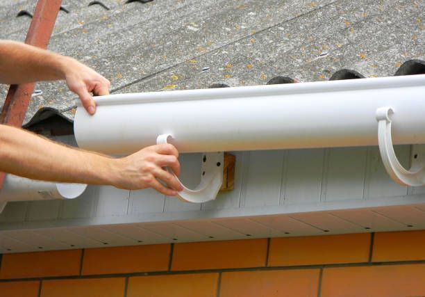 Hands attaching a white gutter to a house, against a roof.