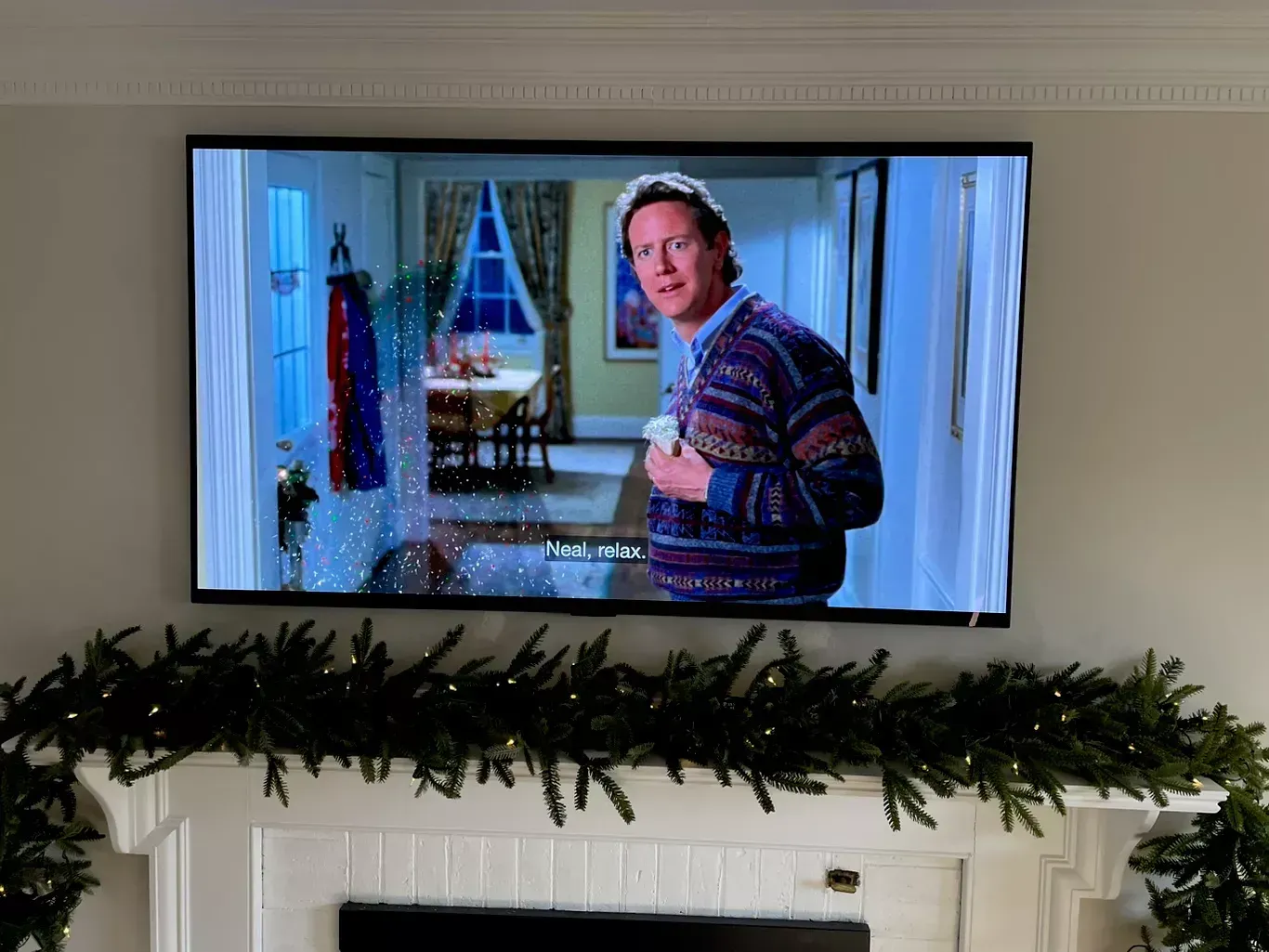 Wall-mounted TV showing a man in a patterned sweater in a bright room, with garland on the mantel below