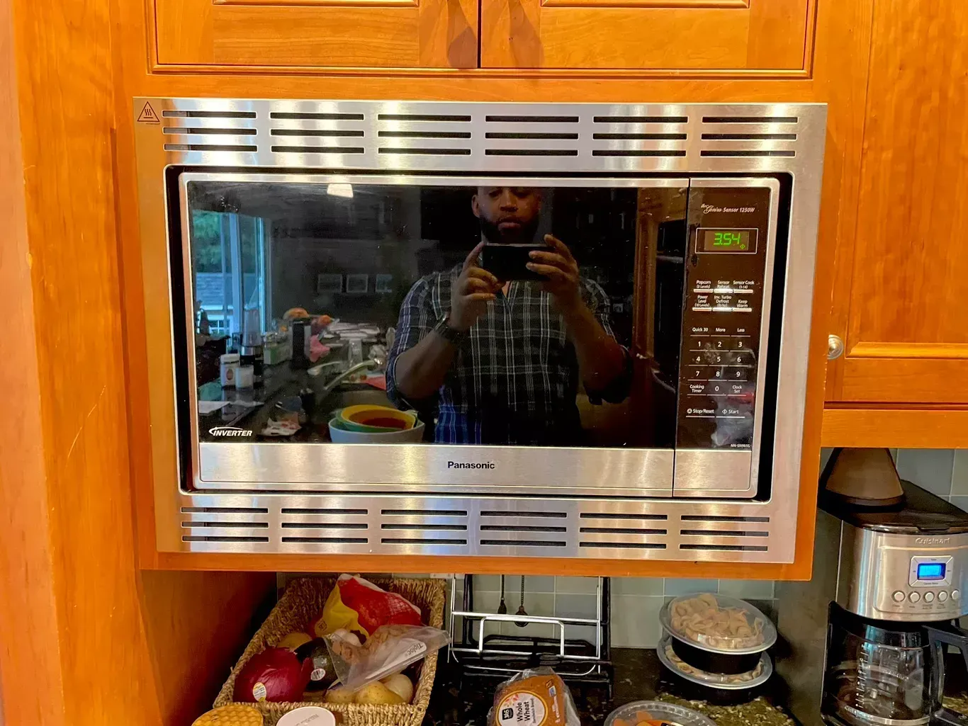 Built-in stainless steel microwave in a wooden kitchen cabinet, reflecting a person holding a phone.