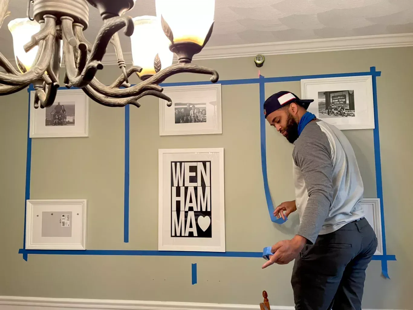 Man measuring a wall with blue painter’s tape around framed pictures under a chandelier.