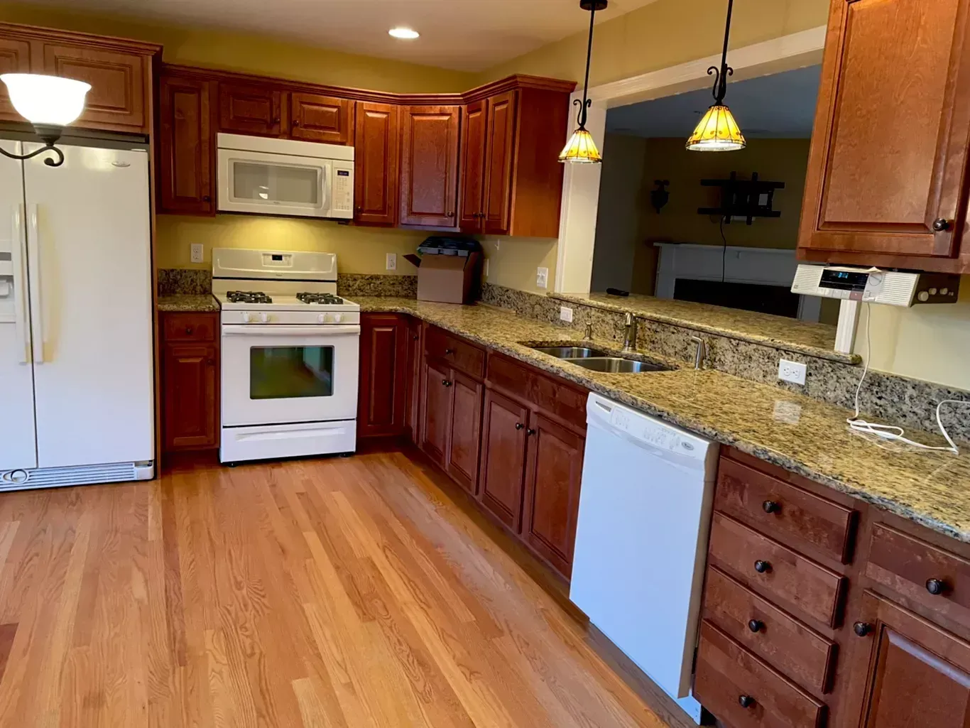 Empty kitchen with wood cabinets, white appliances, granite counters, and pendant lights over a hardwood floor