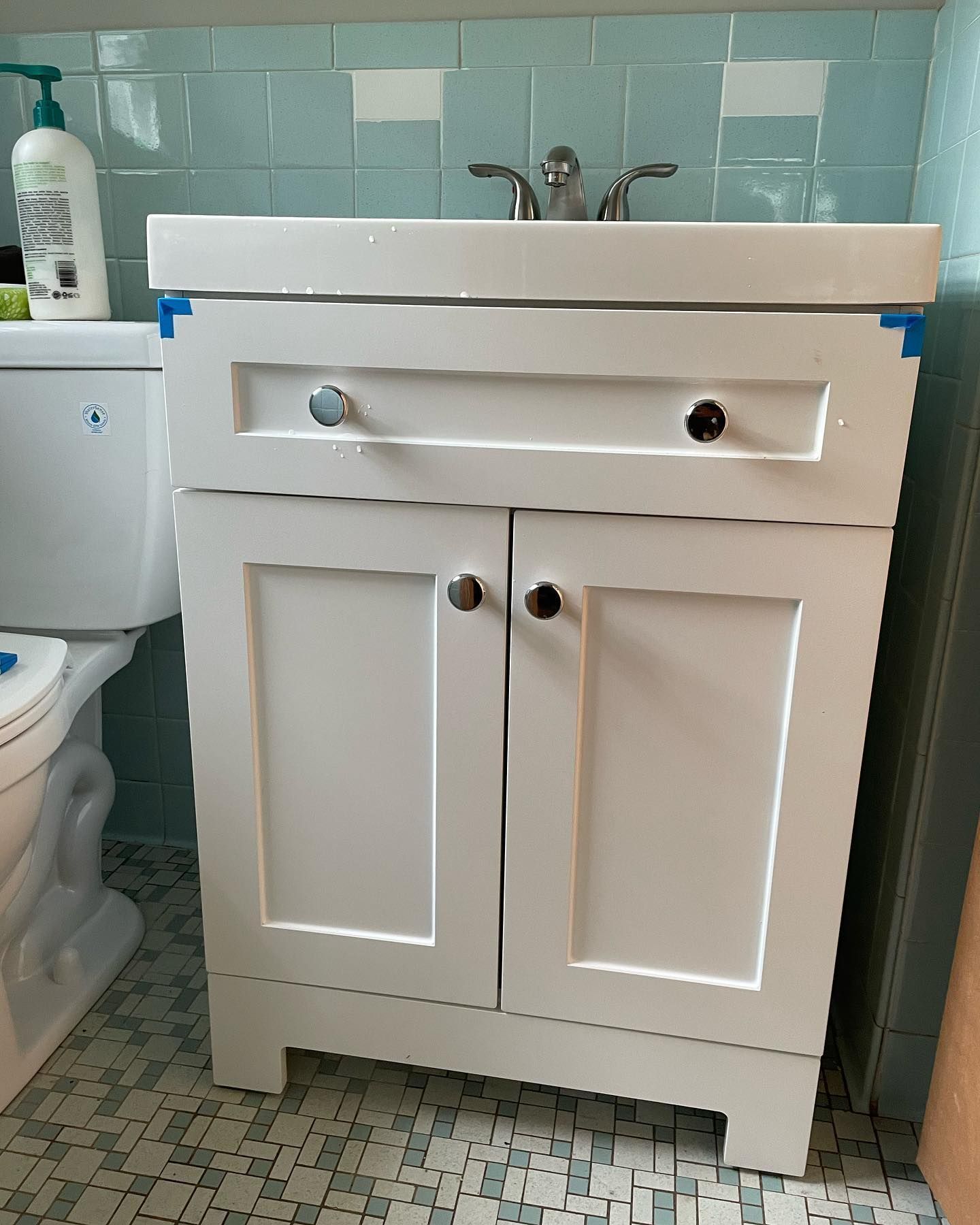 White bathroom vanity with sink and chrome faucet beside a toilet, against light aqua tile walls.