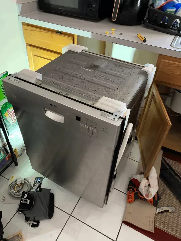 Dishwasher leaning upright in a cluttered kitchen, with a microwave and items on a countertop nearby.