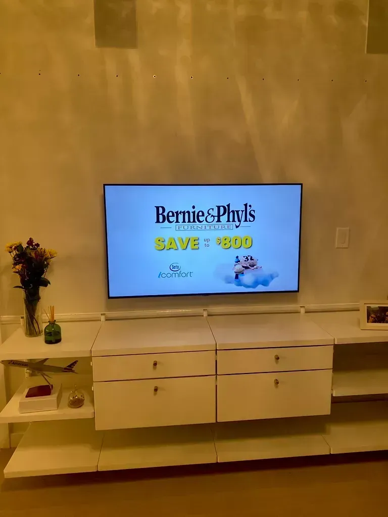 Living room with a TV on a white cabinet displaying a BerniePlus savings promotion.