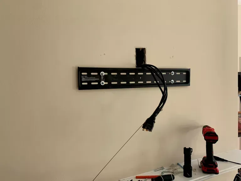 Wall-mounted TV bracket with hanging cables, and a red-and-black drill on a tabletop beneath it.
