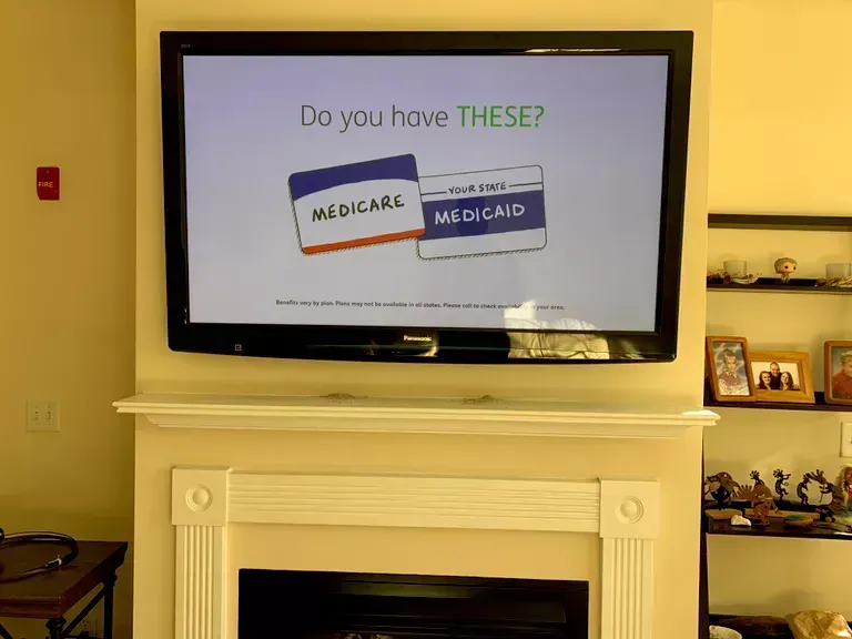 TV above fireplace showing medication ad with two pill bottles on a beige wall