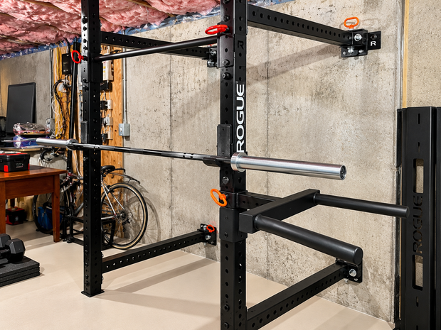 Black Rogue squat rack with safety arms in a basement gym, against a concrete wall.