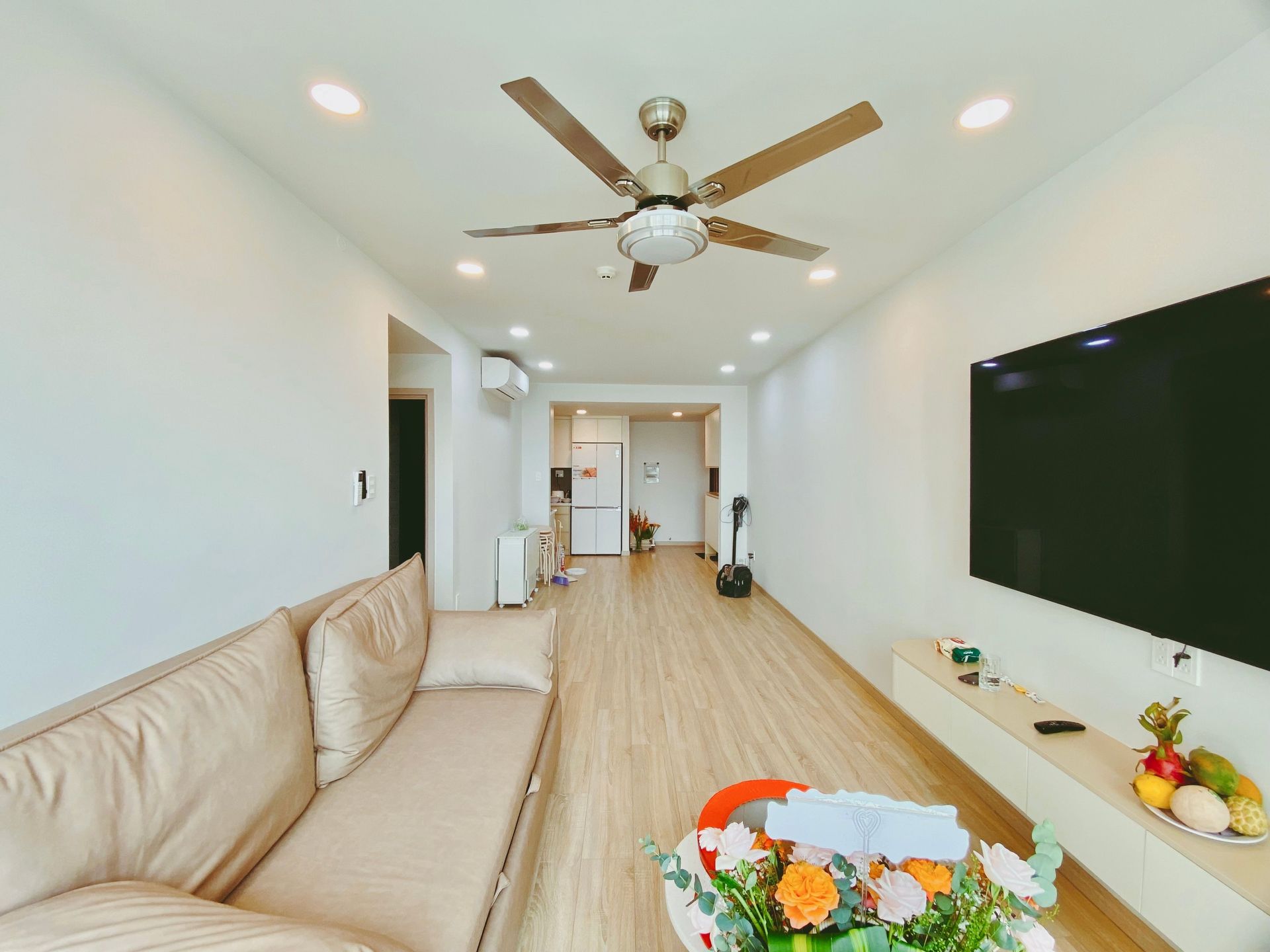 Bright living room with beige sofa, ceiling fan, wall TV, and wood flooring
