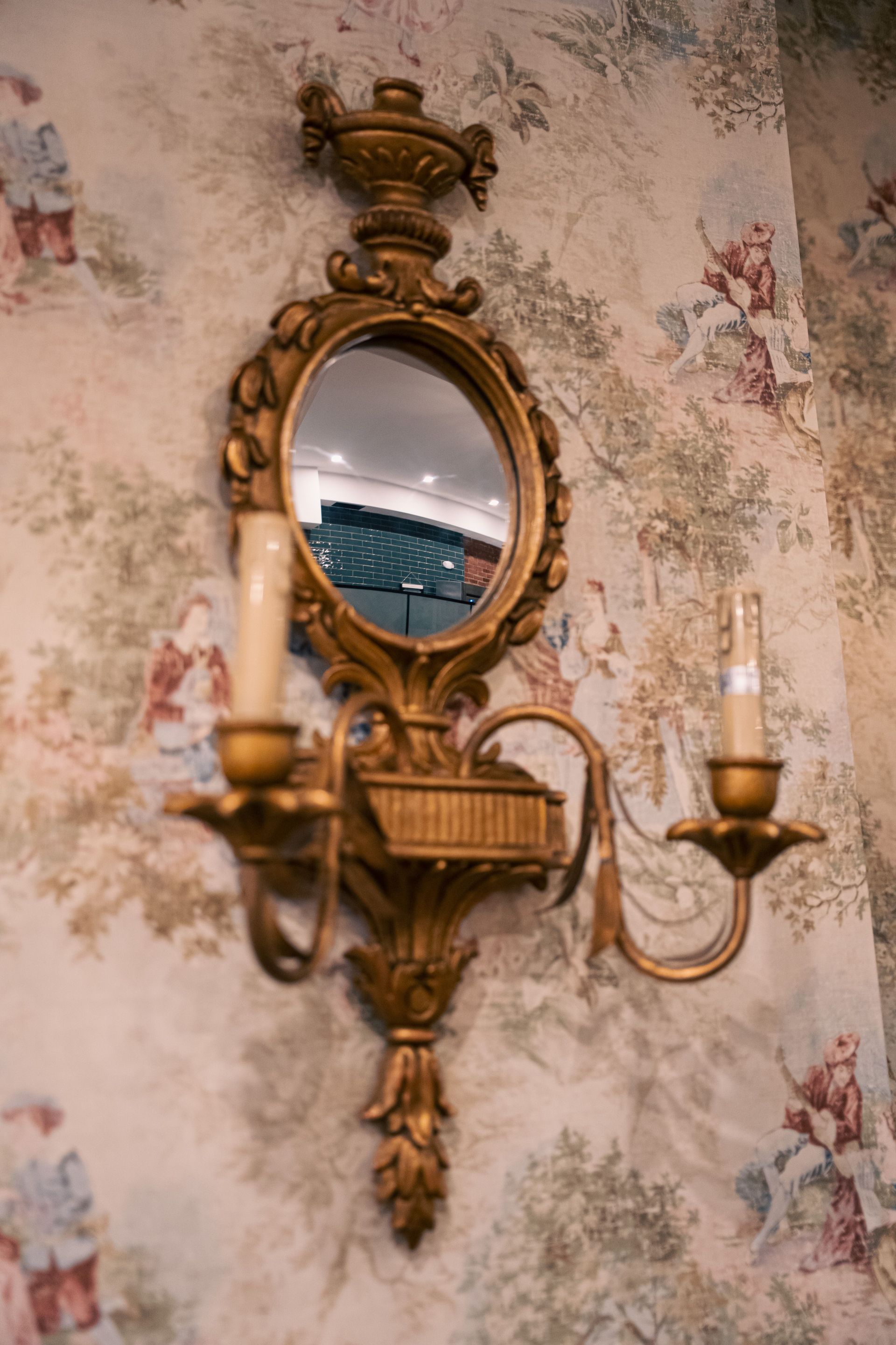 Ornate gold wall mirror sconce with two candles on floral wallpaper, reflecting a room