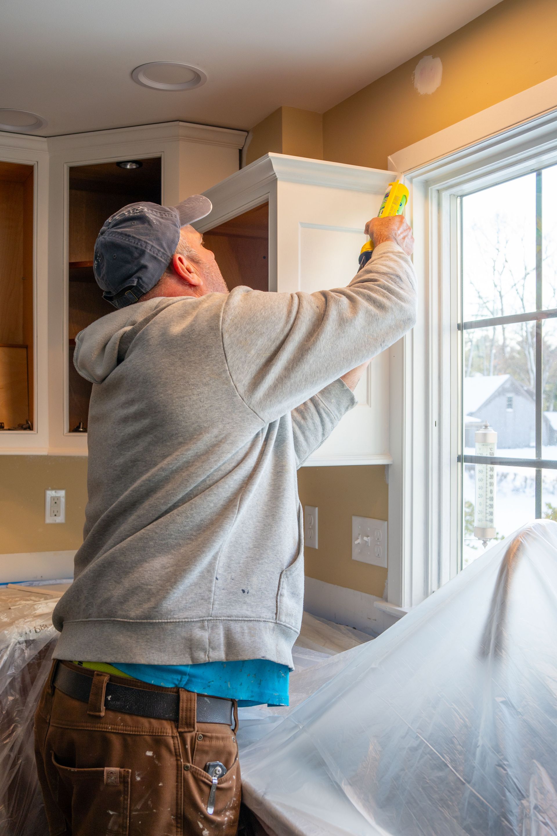 Person painting a kitchen cabinet near a window, with plastic sheeting covering the floor.