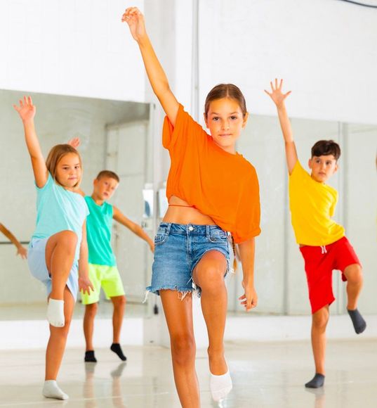 Children in a dance studio practicing moves: arm raised, leg lifted, with happy expressions.