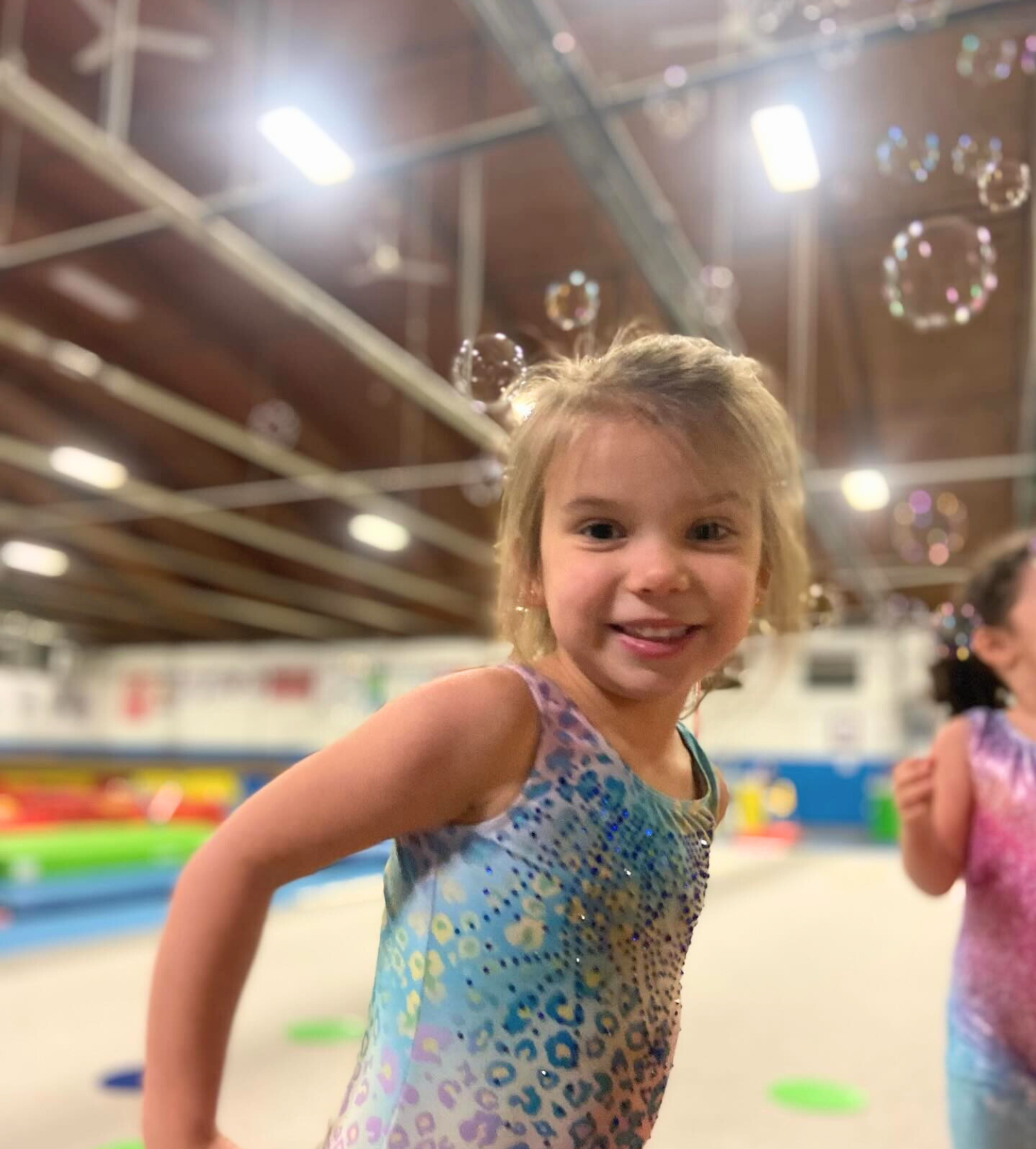 Girl in a blue leotard smiles, surrounded by bubbles, in a gymnastics gym.