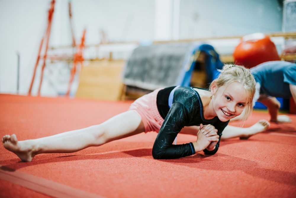 Girl in gymnastics leotard doing the splits on red mat, smiling.