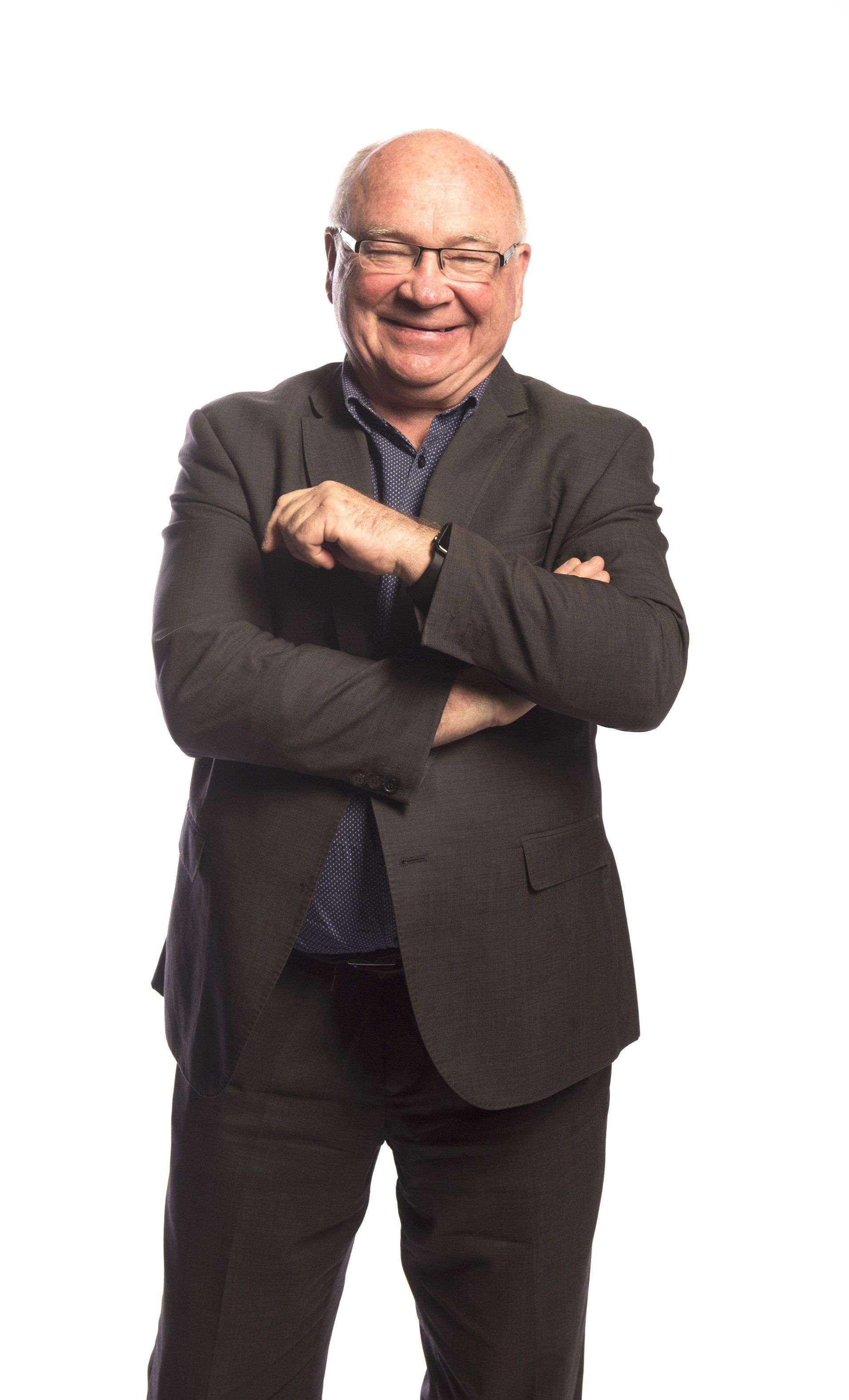 A person wearing glasses and a dark blazer with arms crossed, smiling against a white background.