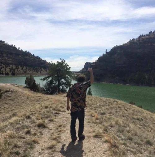 Person standing on a rocky cliff overlooking a turquoise river between forested hills under a cloudy sky