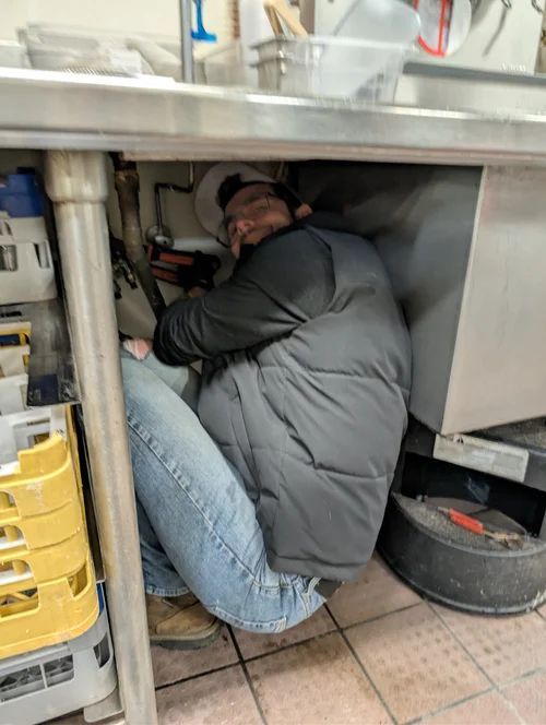 Person crouched under a stainless-steel counter, working in a cramped kitchen space.