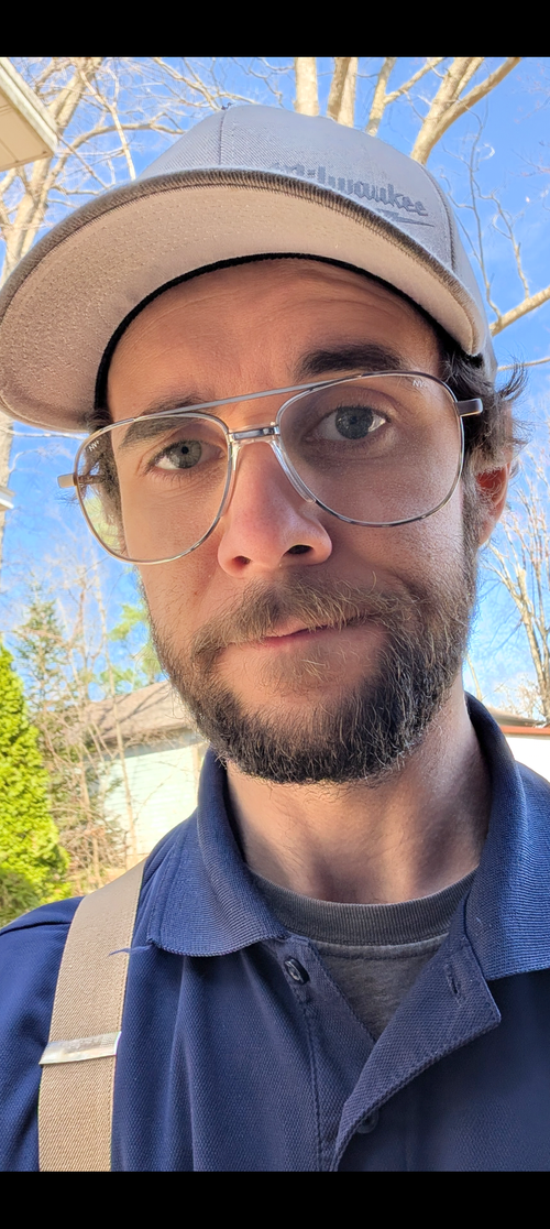 Close-up selfie of a person wearing glasses and a beige cap outdoors under a blue sky.