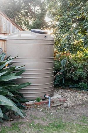 Large beige water tank in a garden, surrounded by plants and gravel.