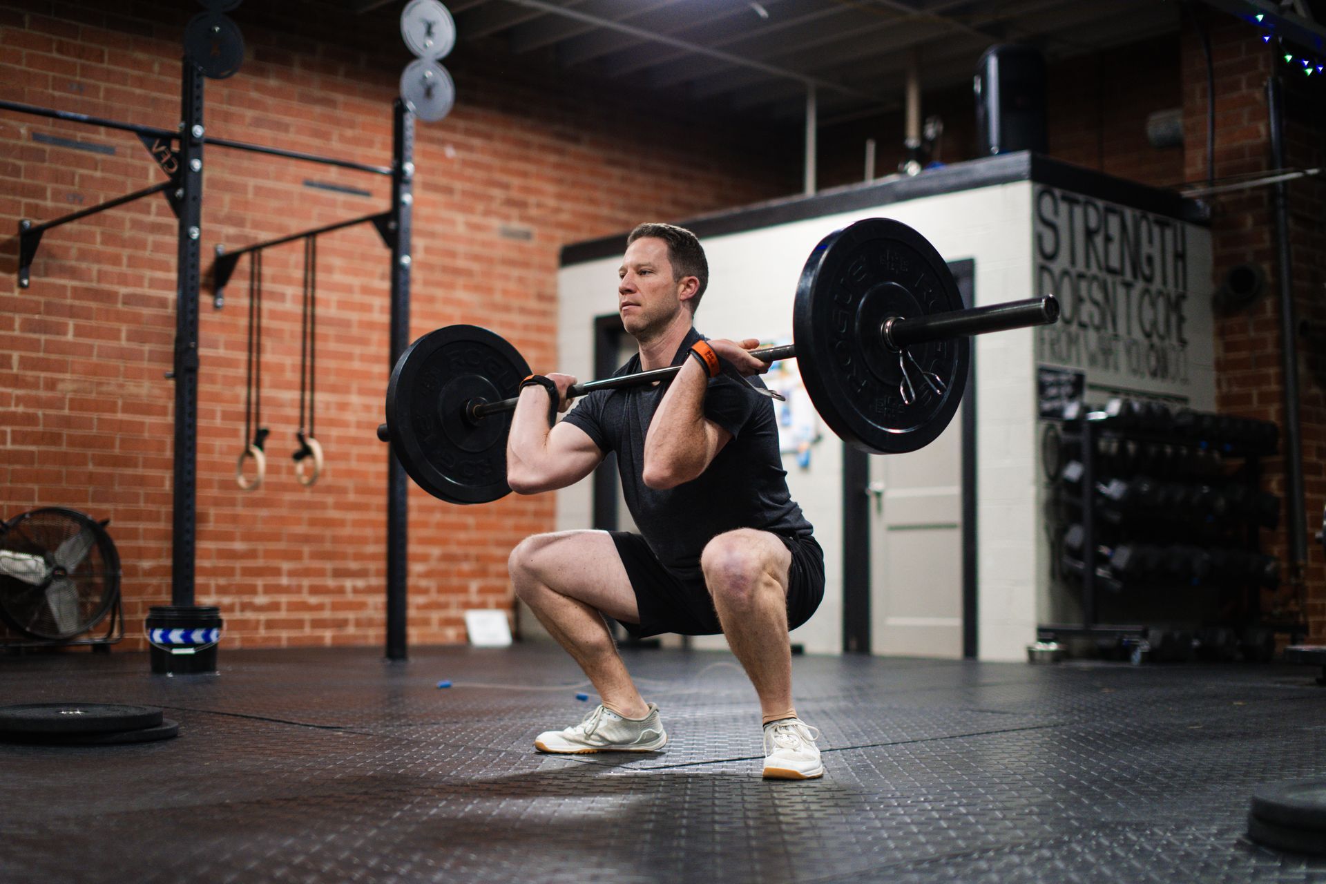 Man in a gym performing a barbell front squat. He is in shorts and a black t-shirt. Brick wall in the background.