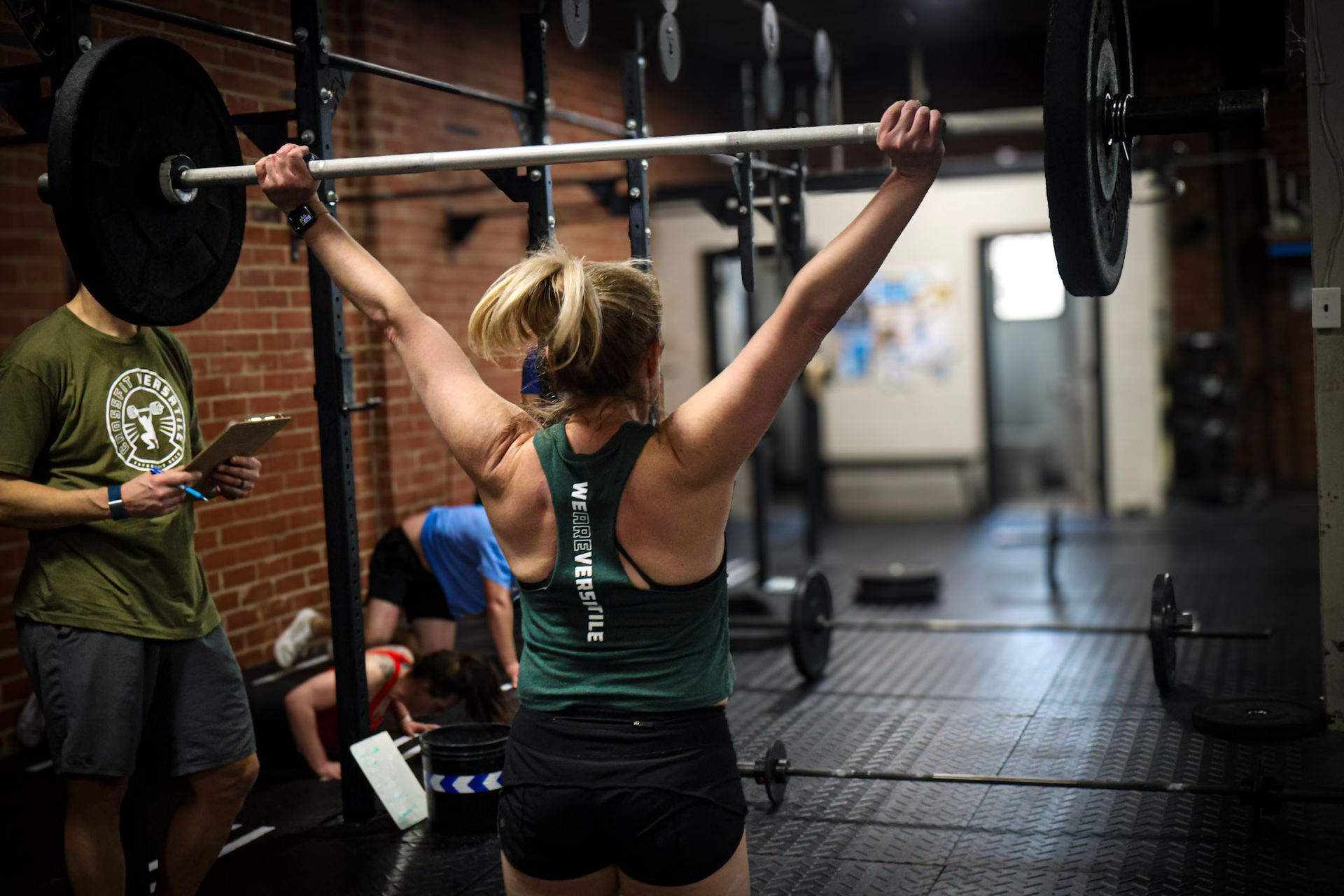 Woman lifting barbell overhead during Olympic weightlifting class in Greensboro NC gym; others nearby, spotter with clipboard.