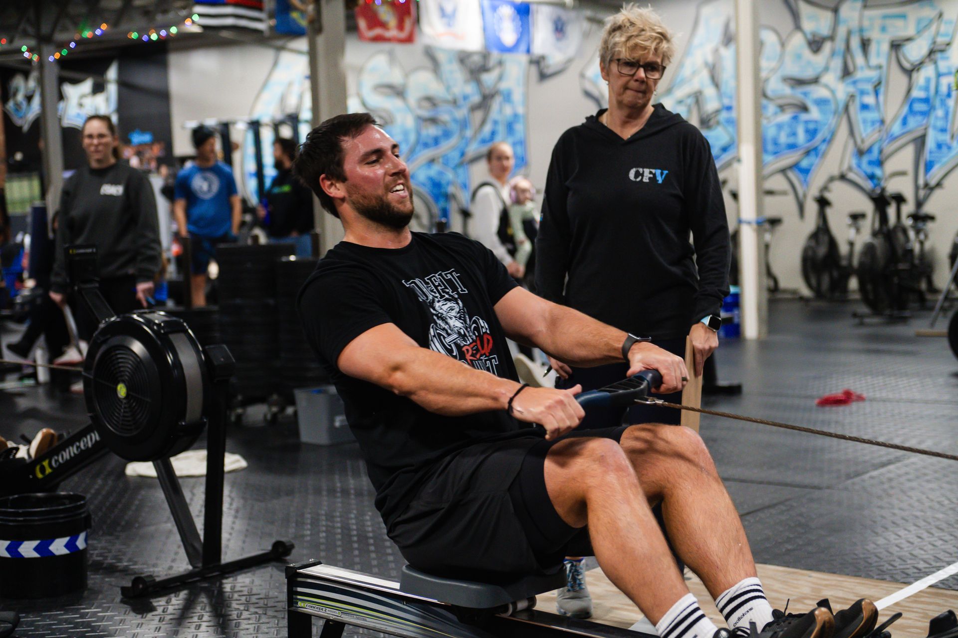 Man rowing indoors, face strained. A CrossFit Versatile instructor watches nearby. Gym setting with graffiti.