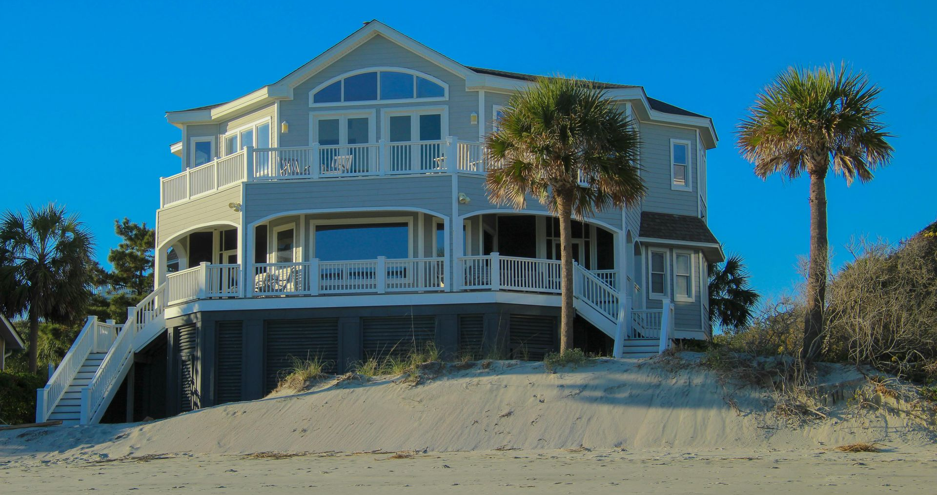 Beachfront house with white siding and a deck, set on sandy dunes under a clear blue sky.