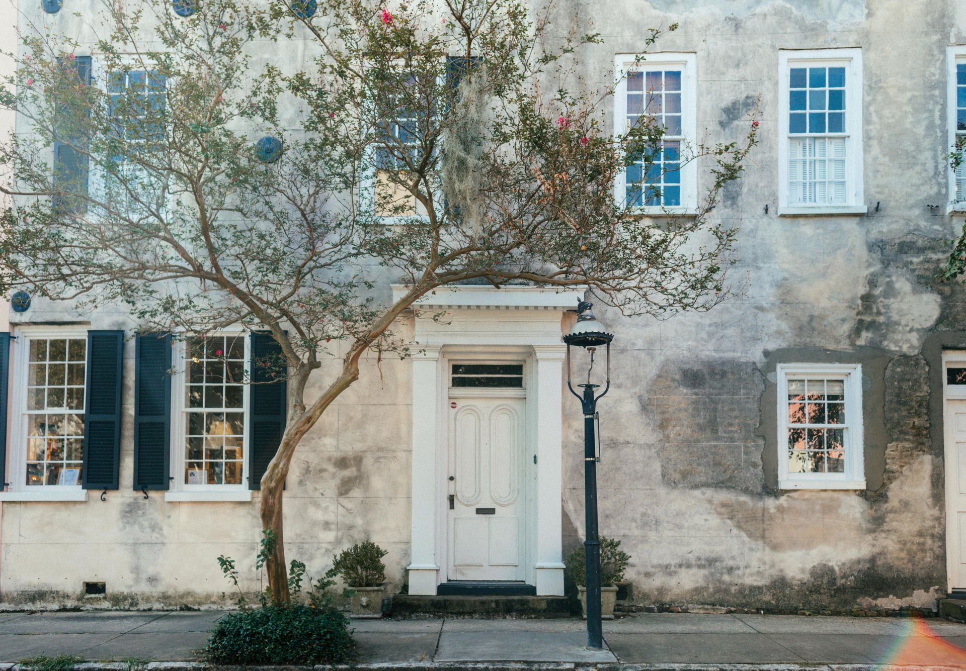 Weathered white townhouse facade with black shutters, a central door, and a streetlamp in front of a tree
