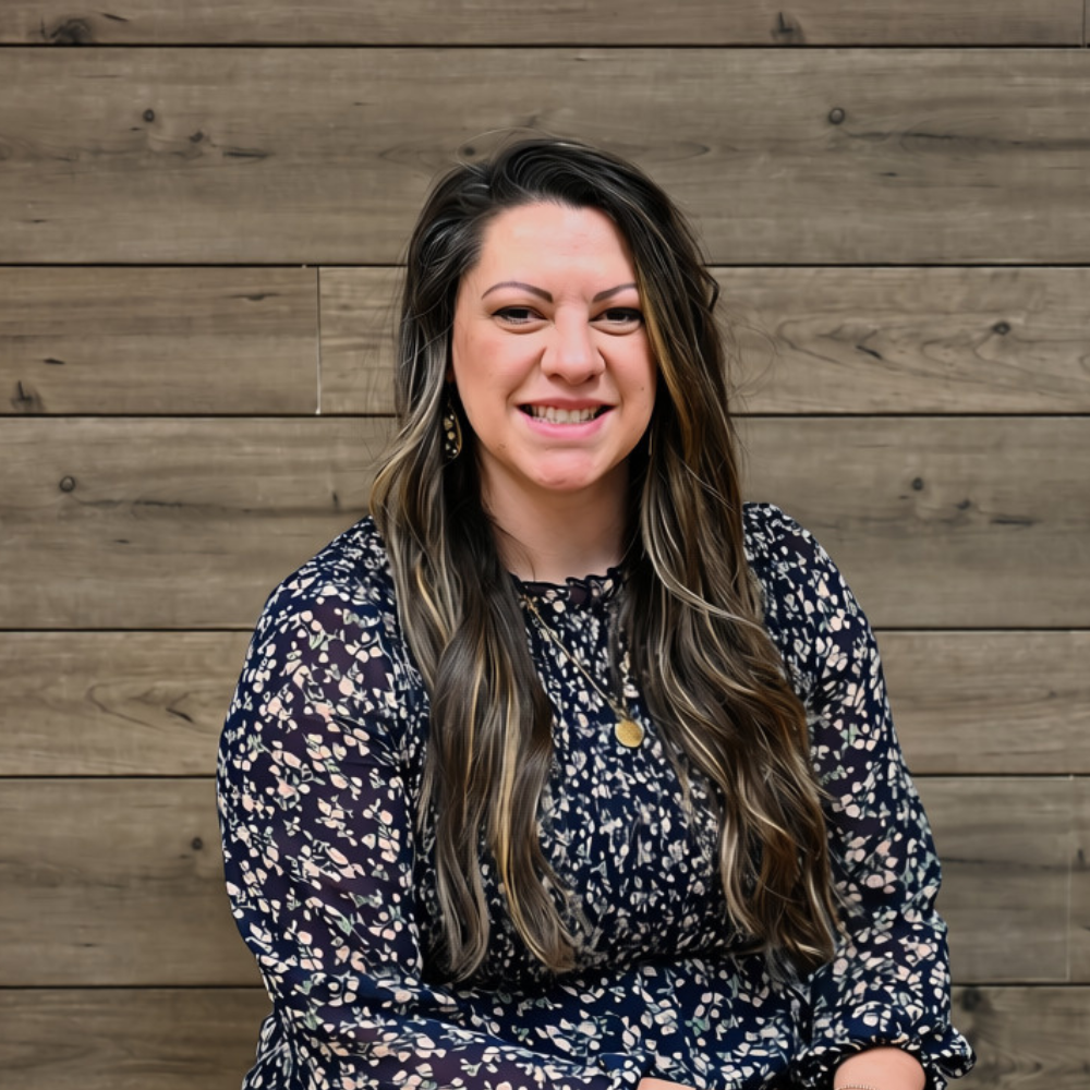 Smiling woman with long dark hair in a navy floral dress against a wooden wall
