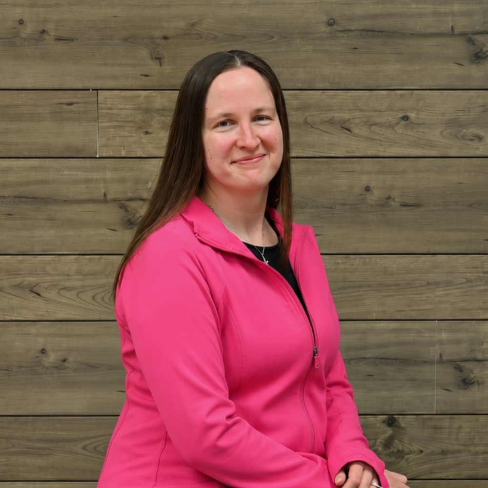 Woman in a bright pink jacket sitting against a wooden wall, smiling at the camera.