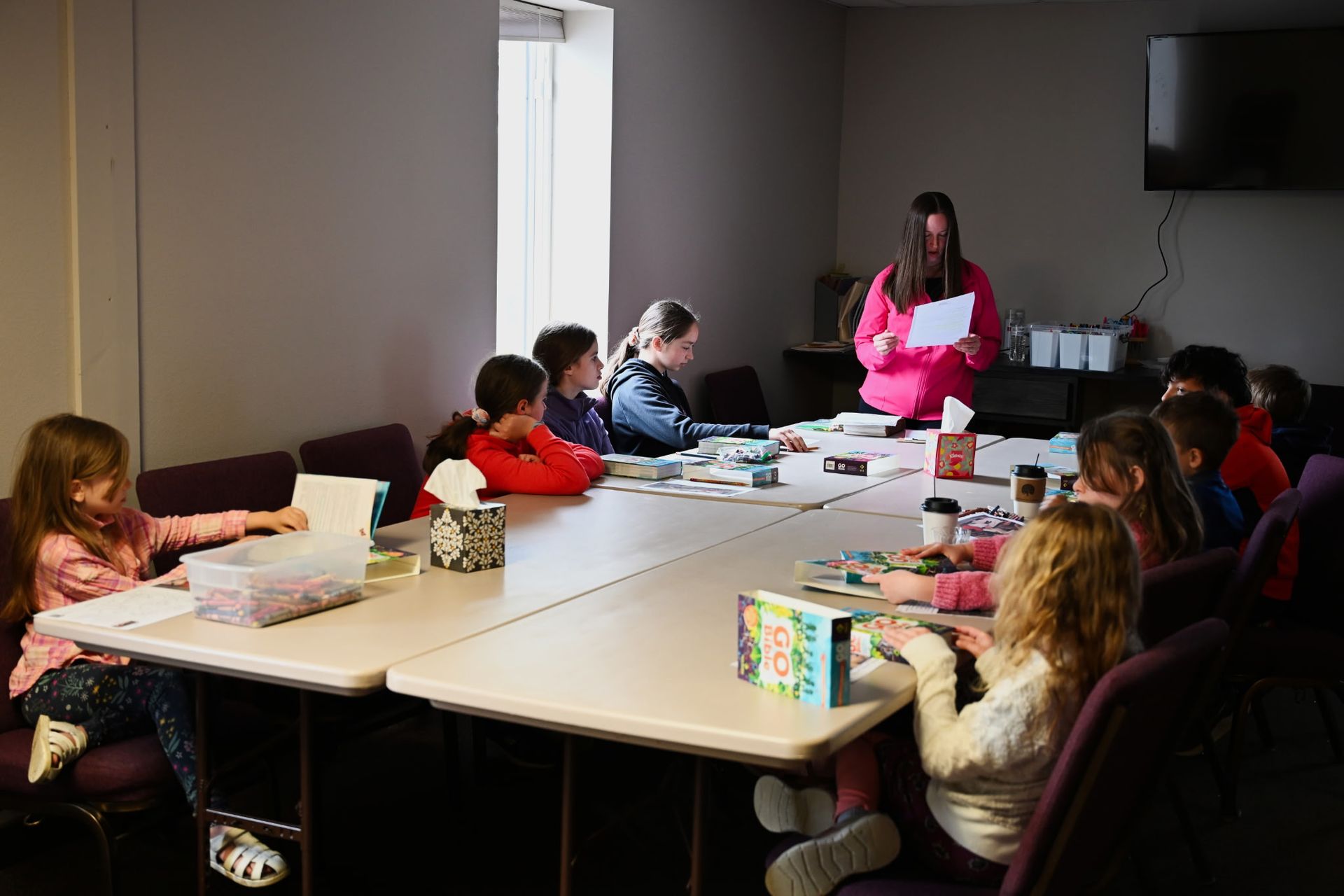 People browsing books at a table in a classroom or library, with one adult assisting two children